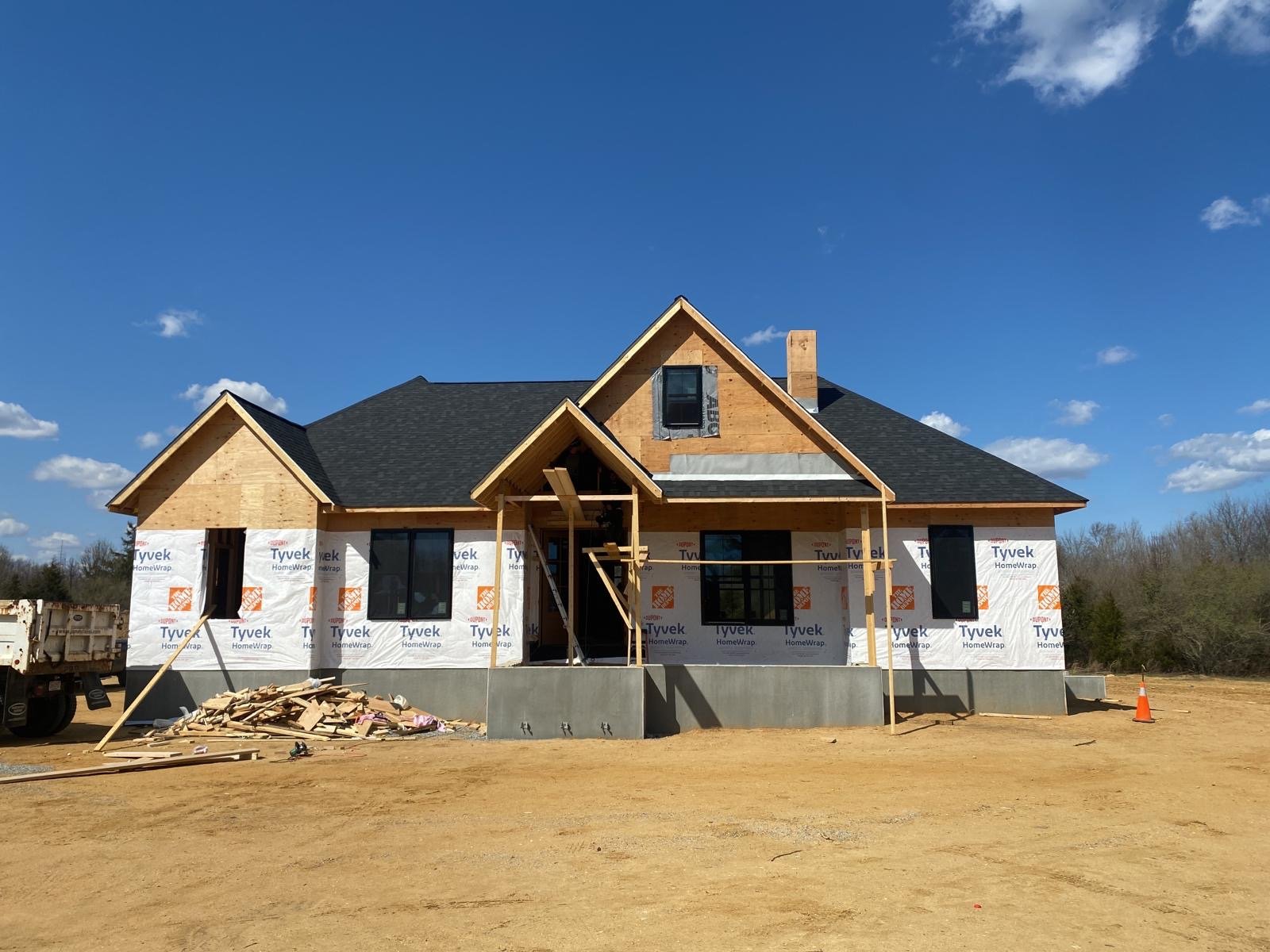 A house under construction with black roof, wooden framing, and white Tyvek vapor barrier on the exterior walls, on a dirt lot under a blue sky.