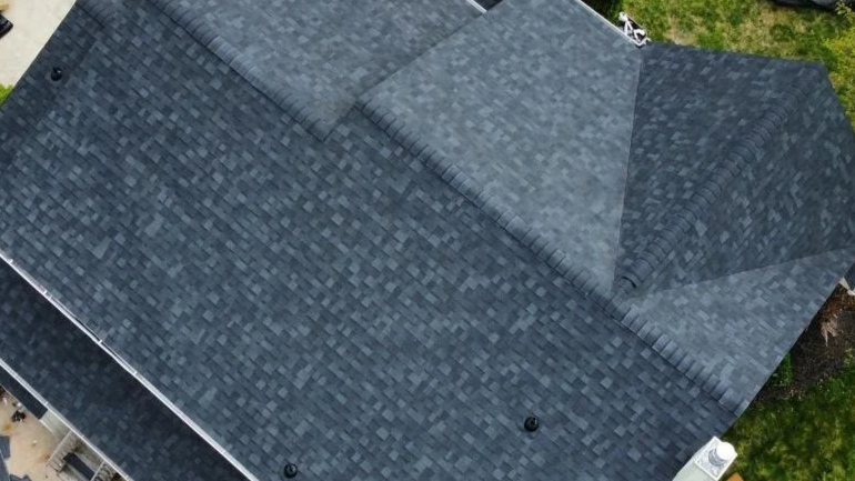 Aerial view of a large house roof with dark gray shingles, showing the peaks and slopes of the roof, and a small section of a backyard with green grass.