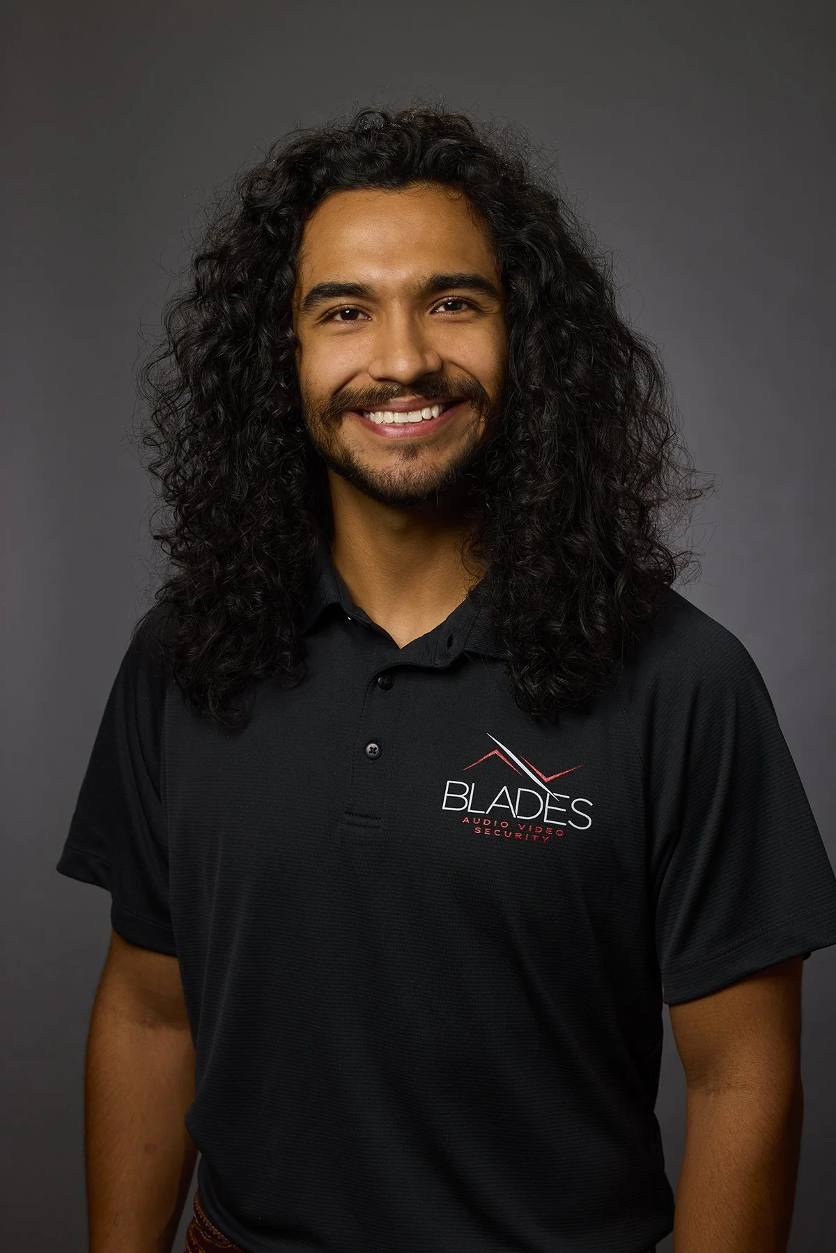 Portrait of a man with long curly hair, smiling, wearing a black polo shirt with 'Blades Audio Video Security' logo, against a gray background.
