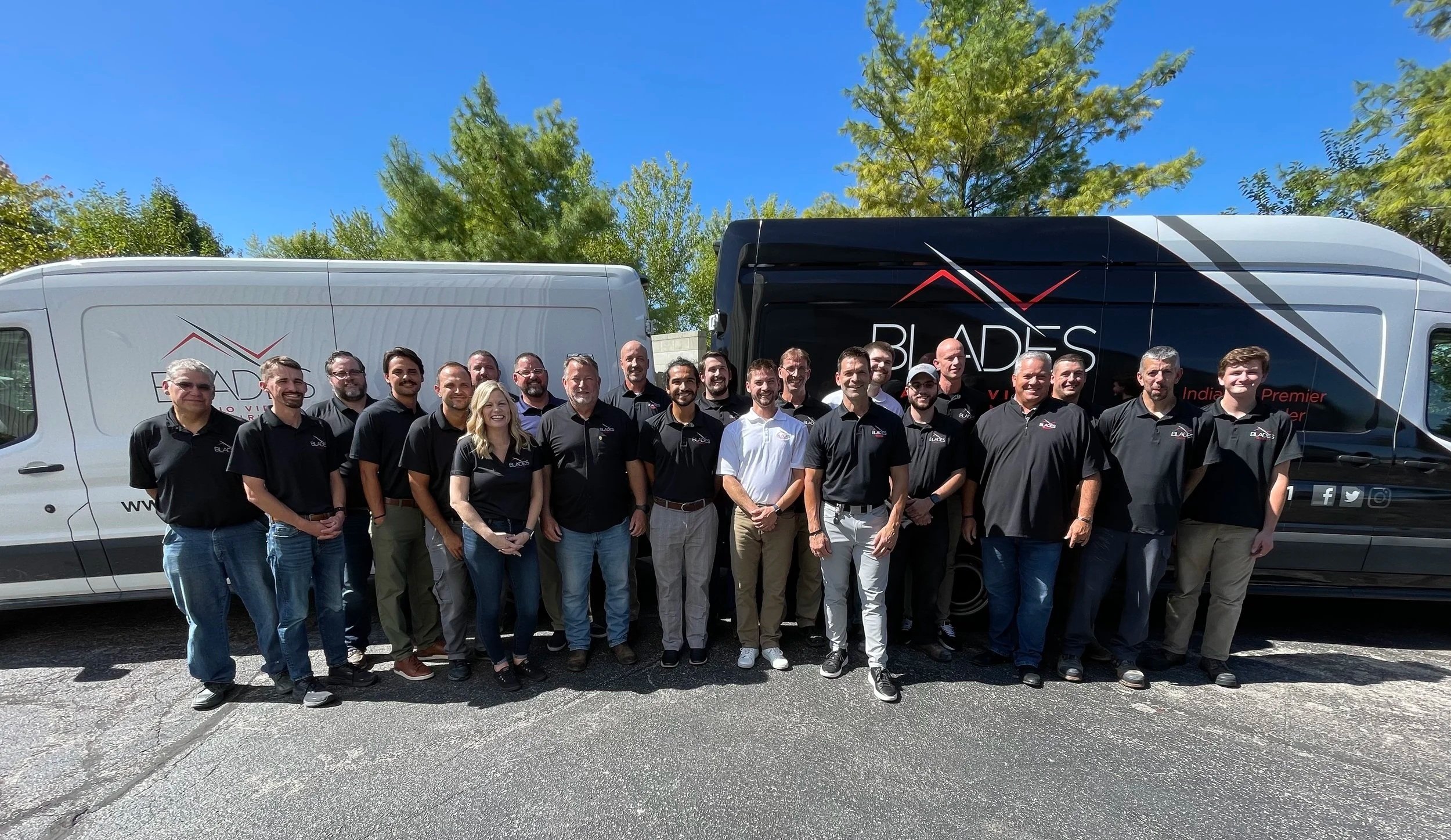 Group of people posing in front of two company vans, outdoor setting with blue sky and green trees.