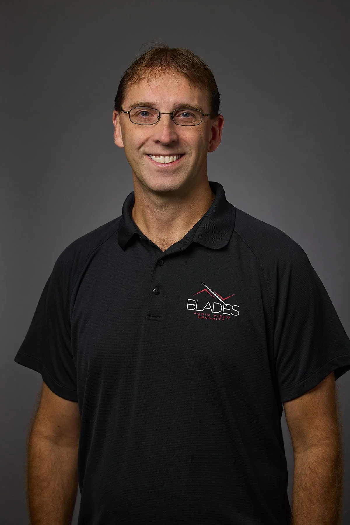 Man wearing glasses and a black polo shirt with 'Blades' logo, smiling, against a gray background.