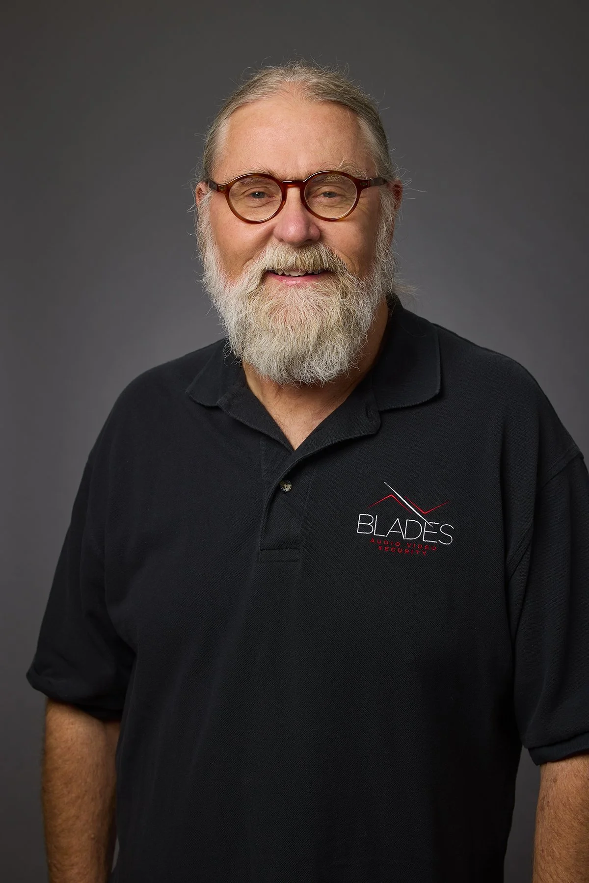A smiling older man with a beard and glasses wearing a black polo shirt with the 'Blades Audio Video Security' logo.