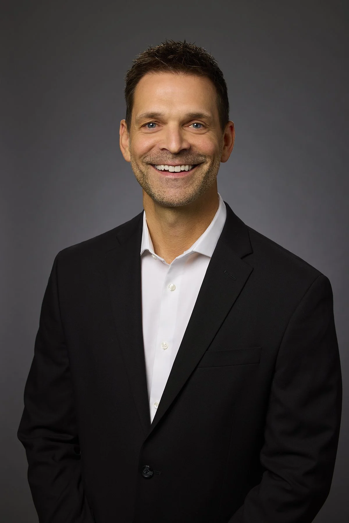 A smiling man with short brown hair, wearing a black suit and white dress shirt, against a dark grey background.