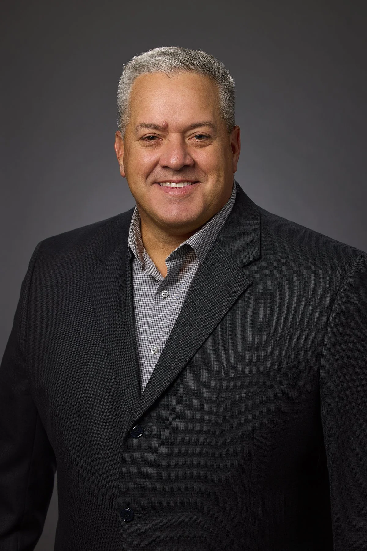A professional man with gray hair, wearing a dark suit and checkered shirt, smiling for a portrait against a gray background.