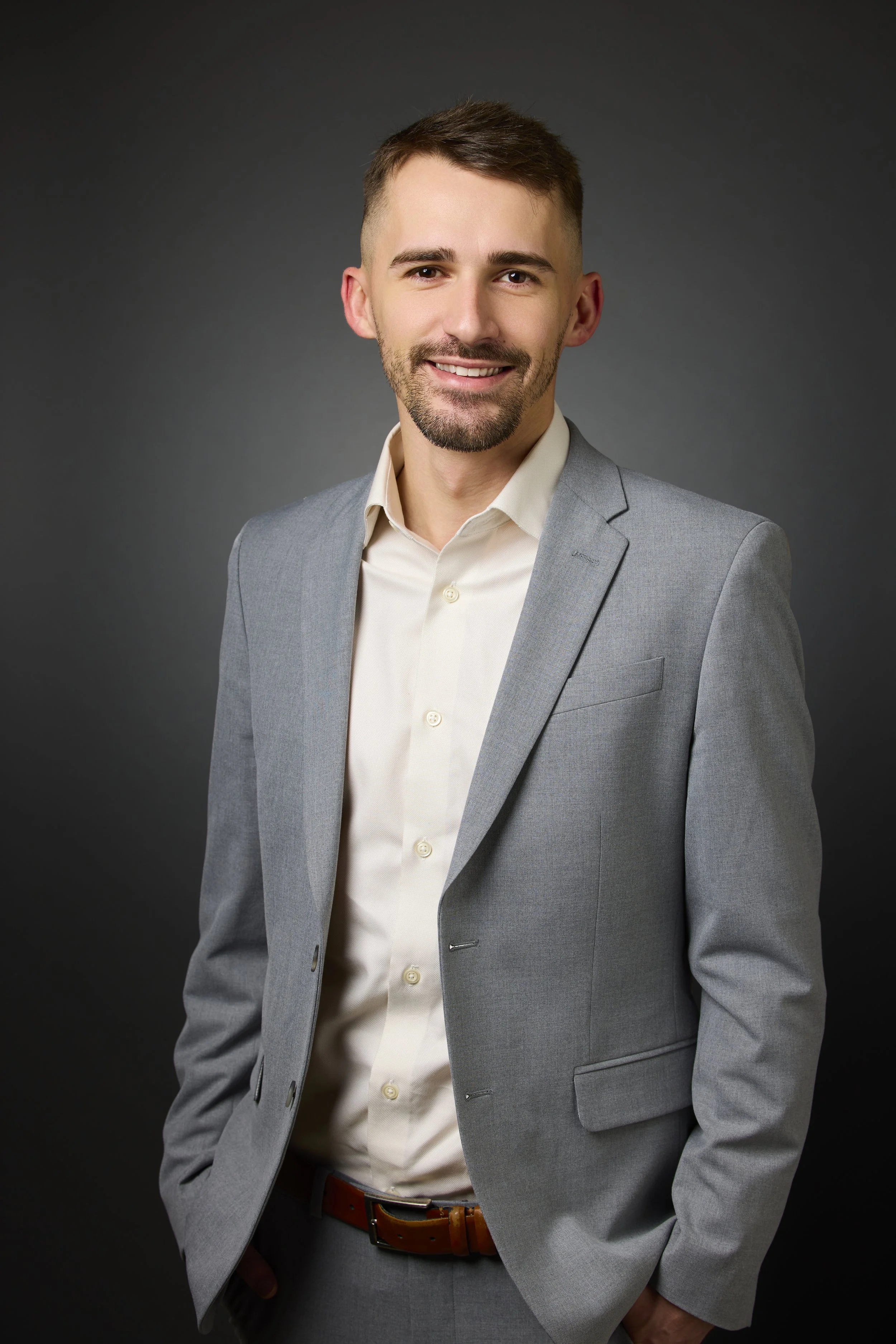A man wearing a light grey suit, smiling against a gray background.