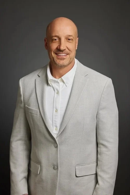 Portrait of a smiling middle-aged man with a bald head and beard, wearing a light grey blazer and white shirt against a dark grey background.