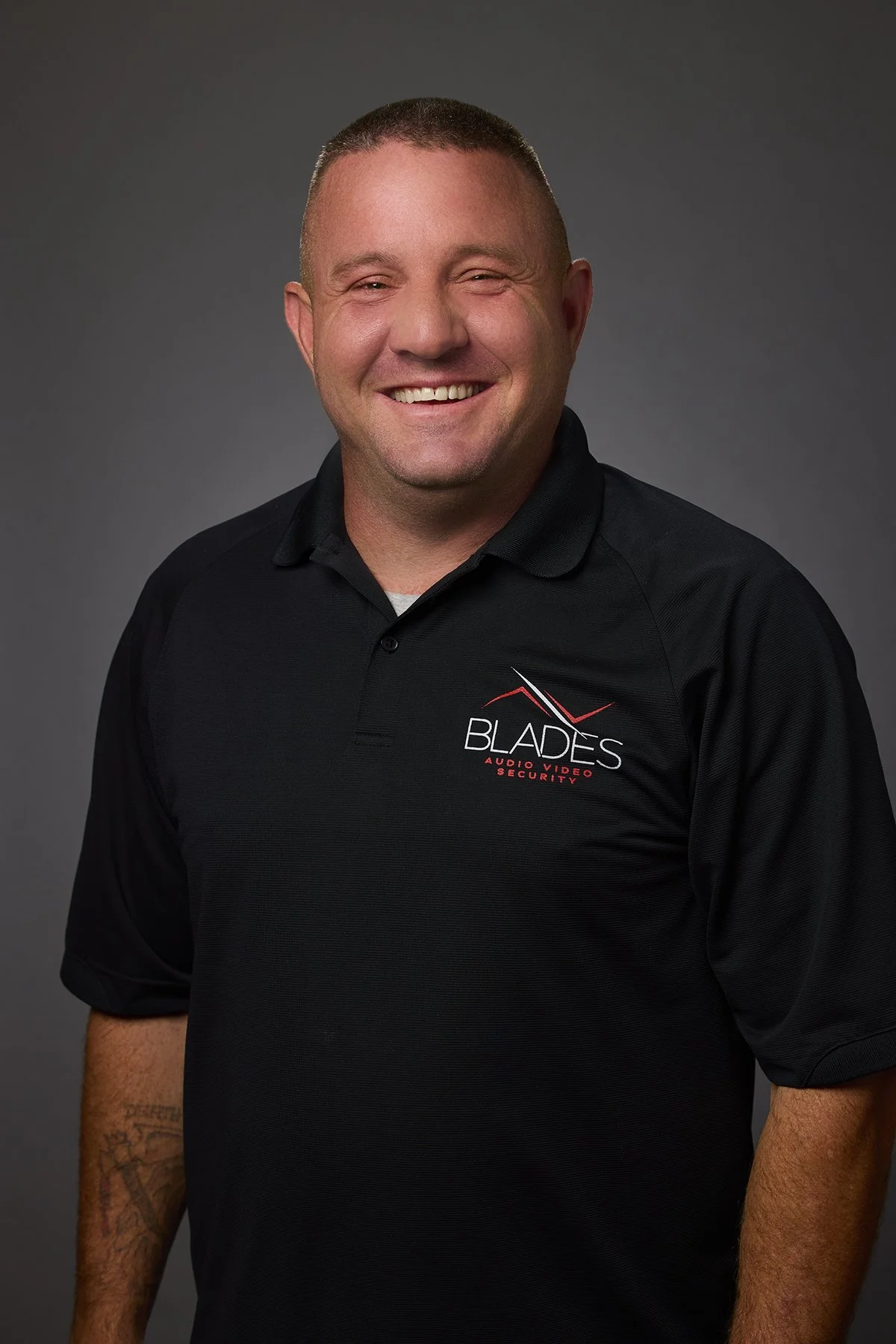 Smiling man wearing a black polo shirt with 'Blades Audio Video Security' logo, standing against gray background.