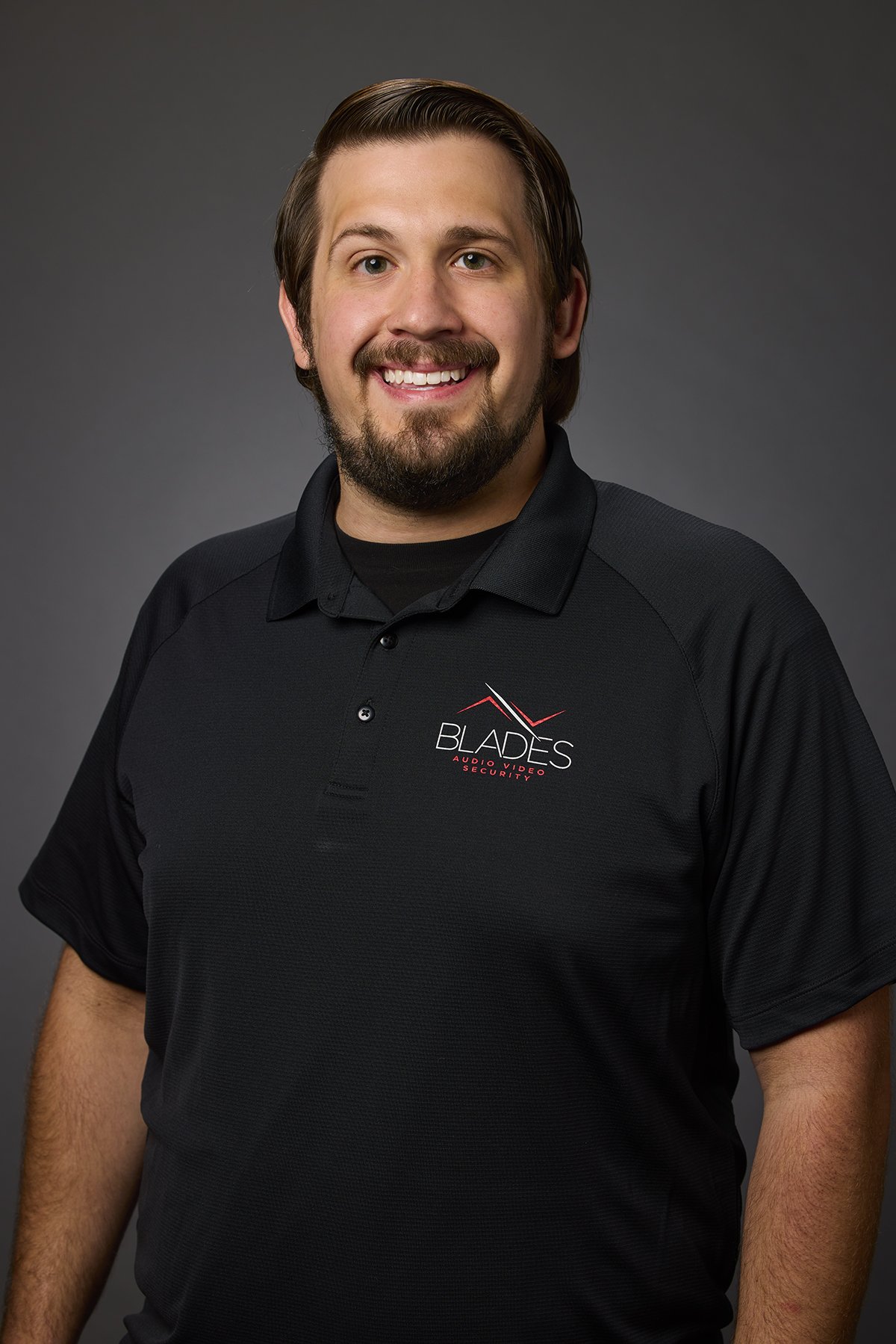 A man with dark hair and a beard smiling, wearing a black polo shirt with a logo that reads 'Blades Audio Video Security' against a gray background.