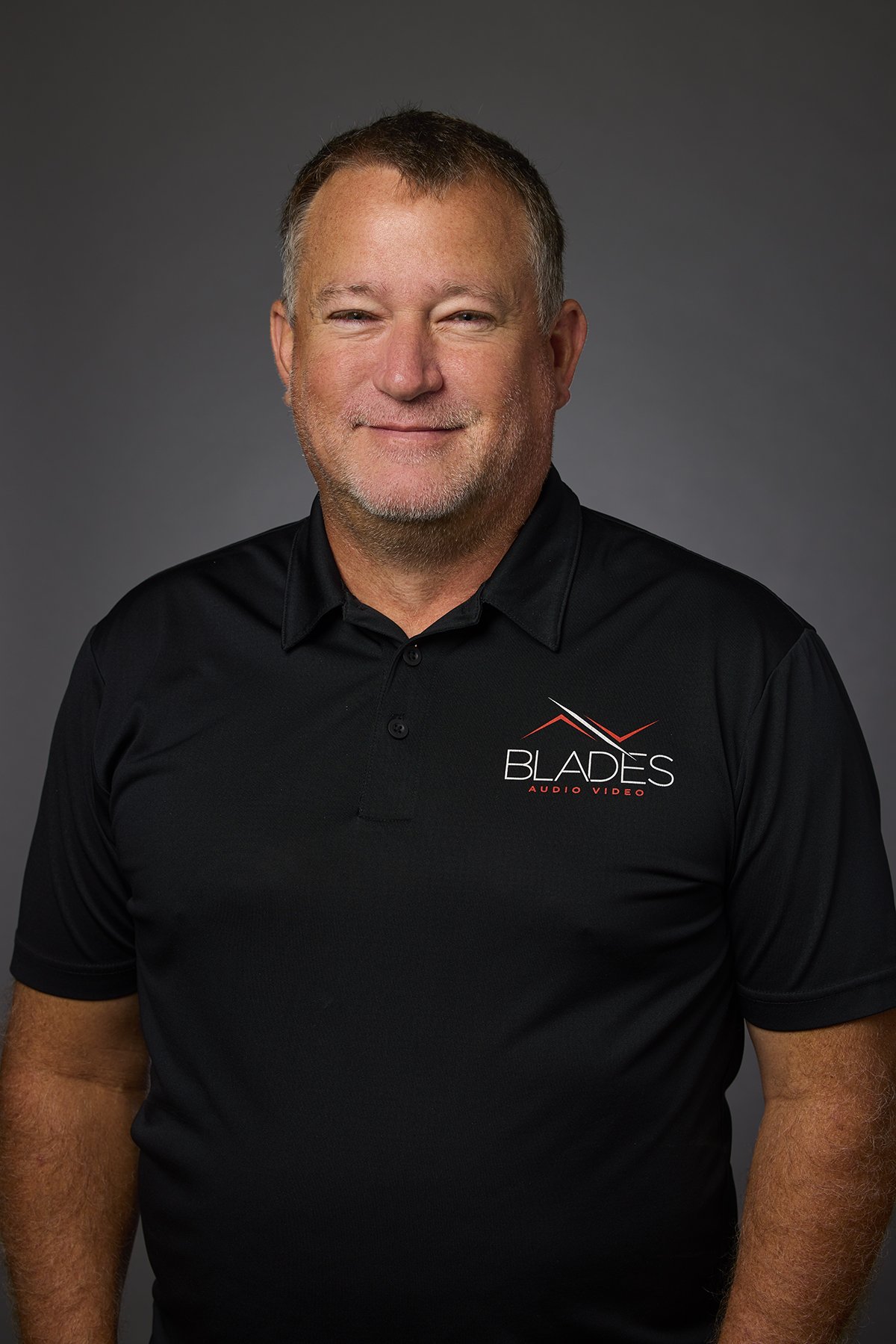 A middle-aged man with light skin, short gray hair, and a beard, wearing a black polo shirt with a "Blades Audio Video" logo, smiling against a gray background.