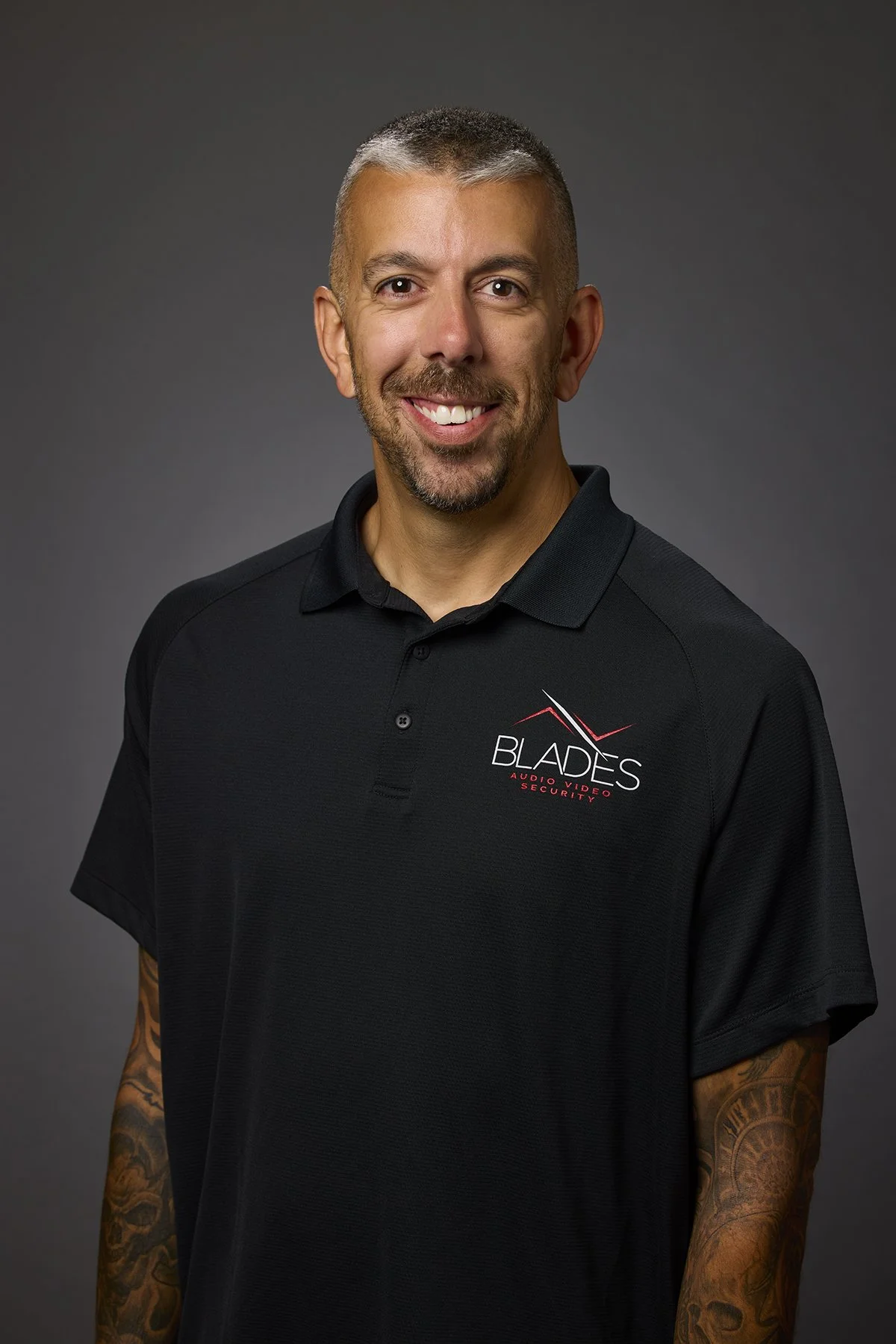Headshot of a man wearing a black polo shirt with 'Blades Audio Video Security' logo, smiling, with tattoos on his right arm, against a dark gray background.