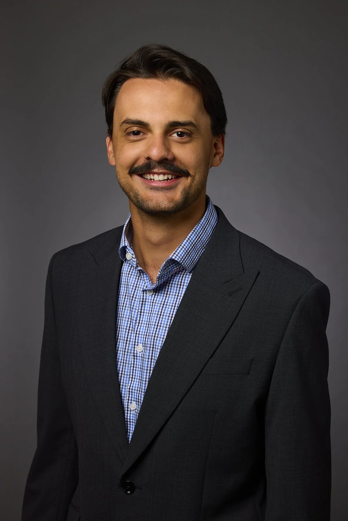 Portrait of a smiling man in a dark suit and checkered shirt against a gray background.