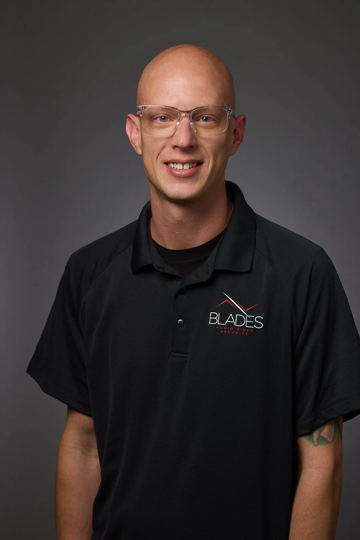 Portrait of a man with glasses, bald head, smiling, wearing a black polo shirt with a logo that says 'Blades Audio/Video Security'.