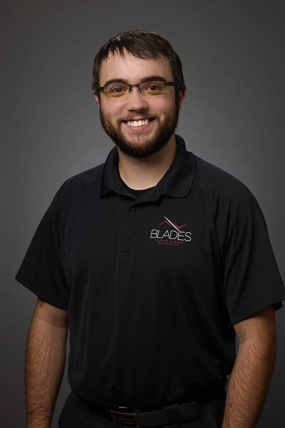 A smiling man wearing glasses and a black polo shirt with 'Blades Audio Video Security' logo, standing against a gray background.