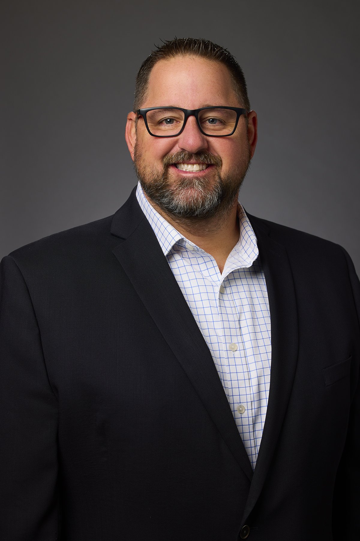 Professional headshot of a man with glasses, beard, and short hair, wearing a black suit jacket and a checked white shirt.