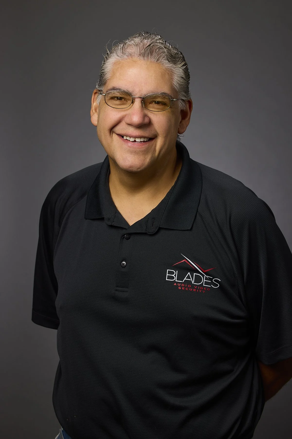 Man with gray hair and glasses smiling, wearing a black polo shirt with a logo that reads 'Blades Audio Video Security'.