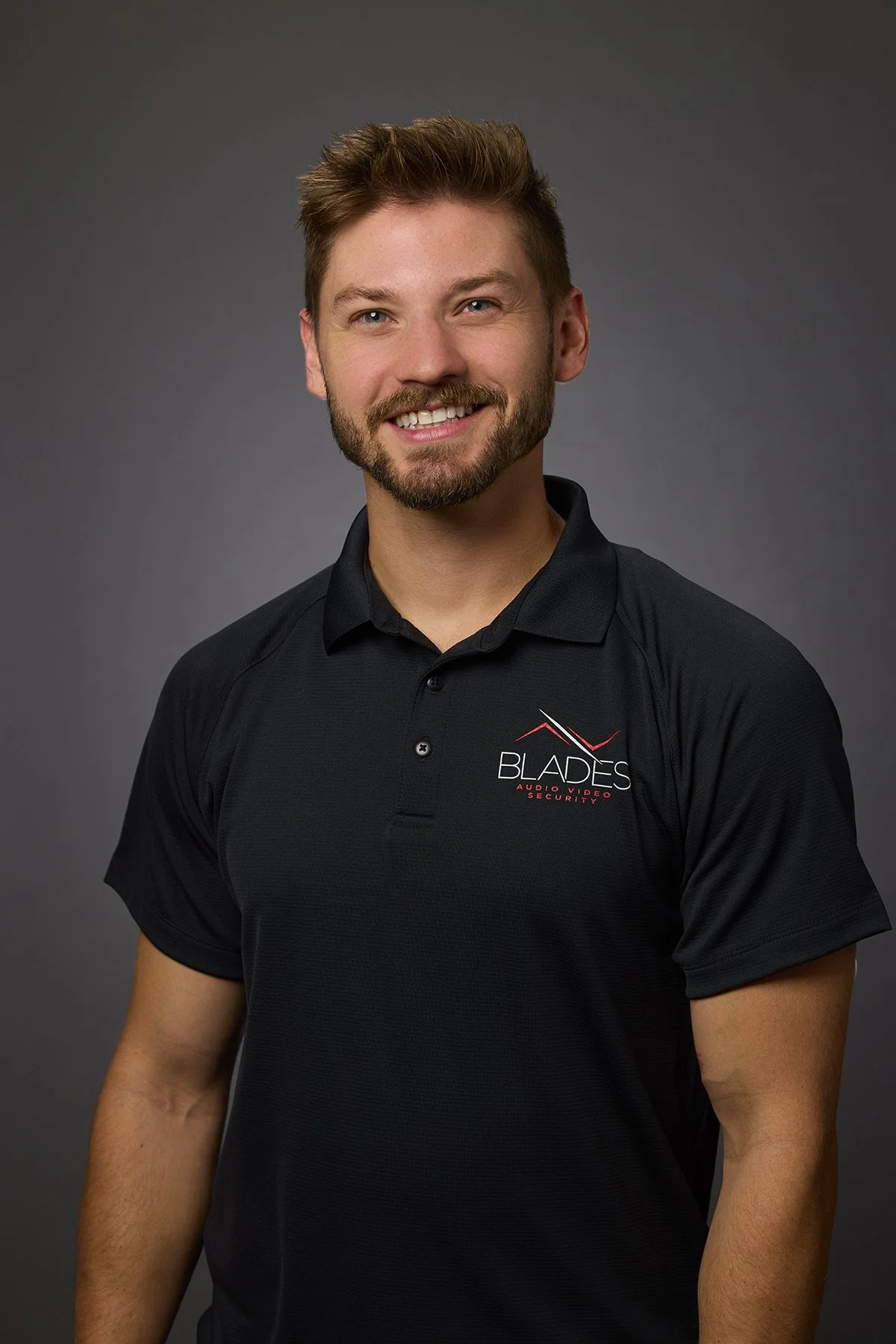 Portrait of a smiling man with brown hair and a beard, wearing a black polo shirt with a company logo that says 'Blades Audio Video Security' on the chest, against a gray background.