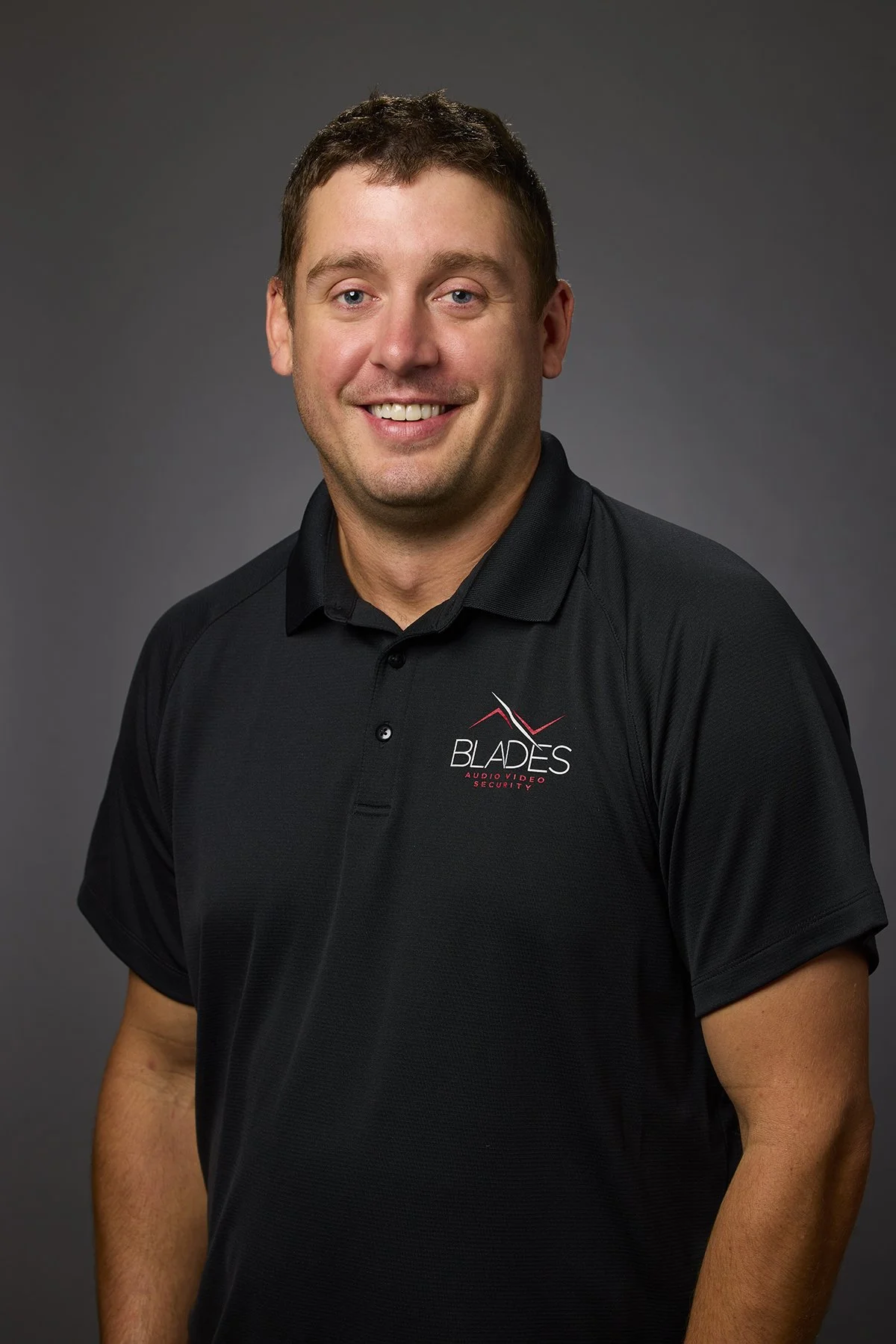 A smiling man wearing a black polo shirt with a logo that reads 'Blades Audio Video Security' against a gray background.