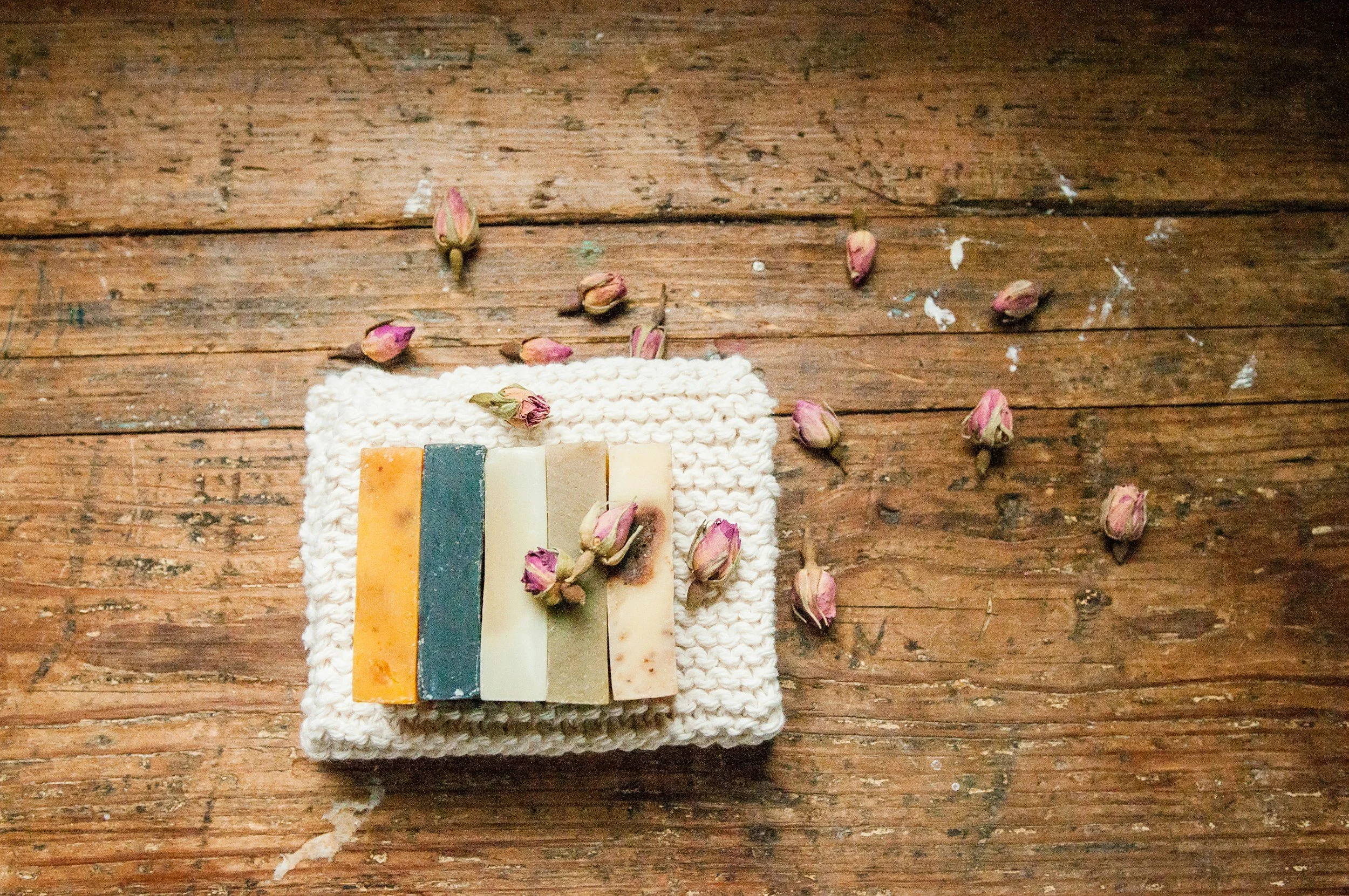 A stack of five colorful soap bars on a white knitted cloth, surrounded by pink dried rosebuds, on a rustic wooden surface.