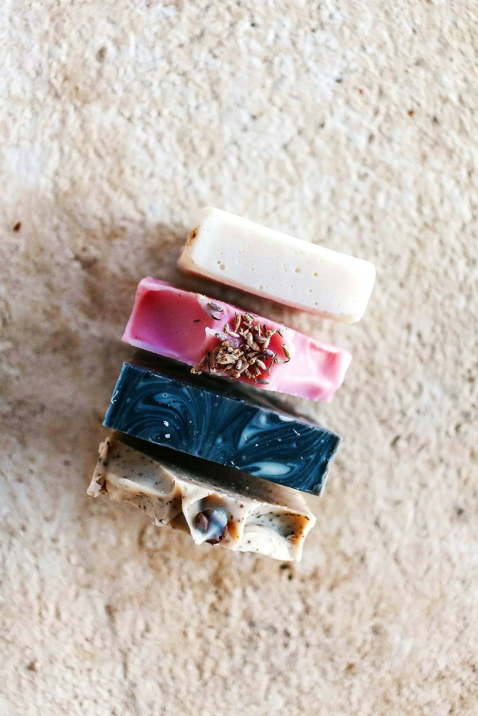 A vertical stack of four different types of soap bars on a sandy surface. The top soap is white, the second is pink with dried lavender on top, the third is dark blue with white swirls, and the bottom soap is beige with mixed inclusions.