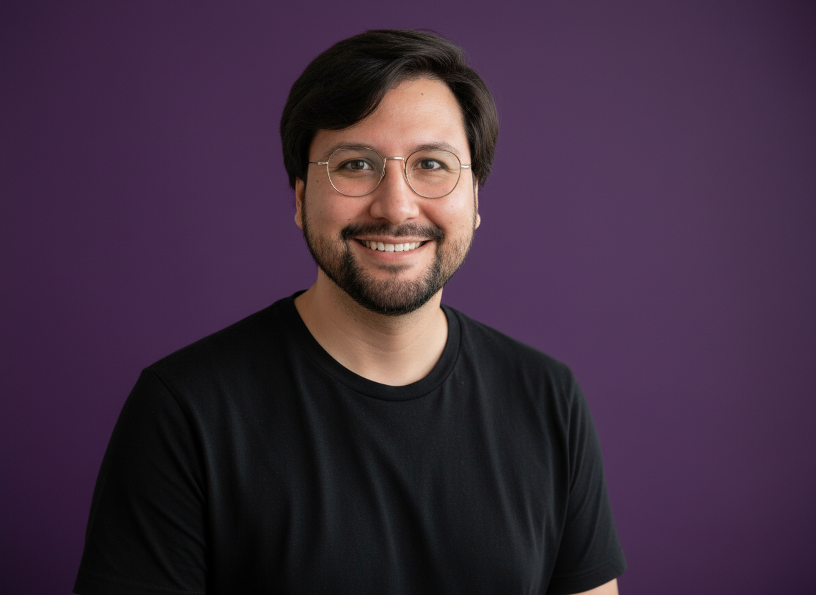 A smiling man with glasses and a beard, wearing a black t-shirt, standing against a purple background.
