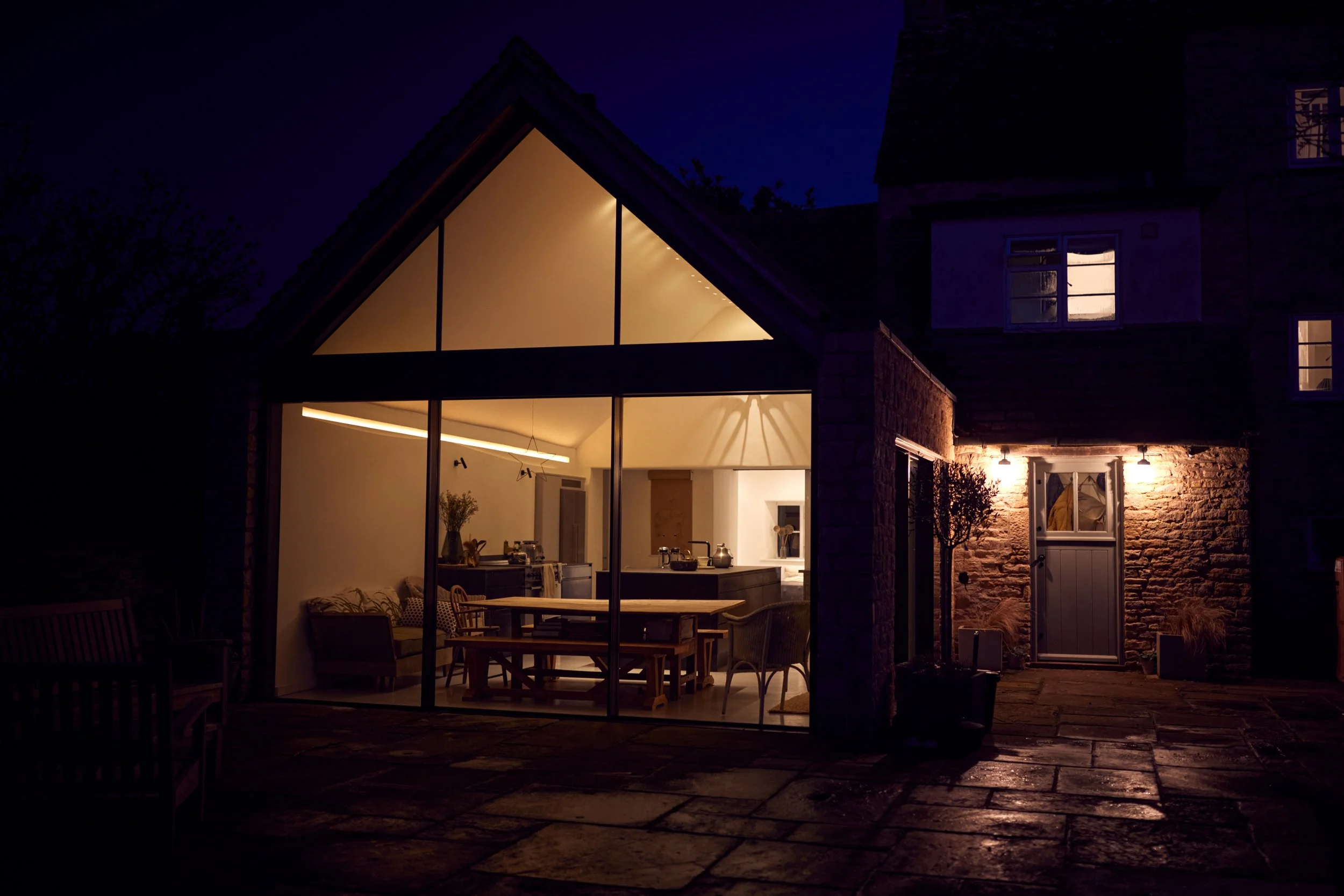 A house with a illuminated modern interior visible through large glass windows at night, featuring a dining area and kitchen, with a brick exterior and small lights outside.