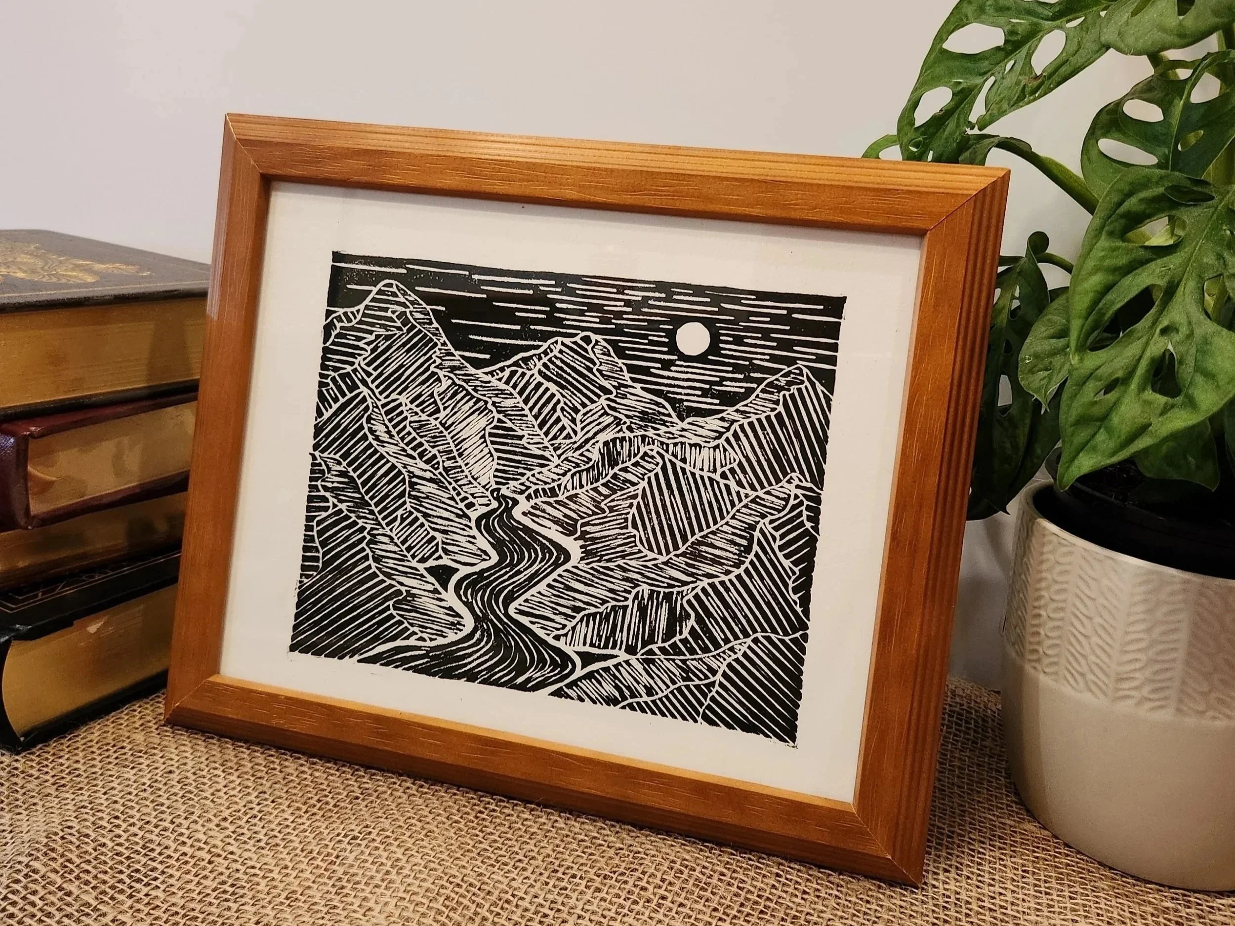 Black and white framed artwork of a mountain landscape with a river and moon, placed on a textured surface next to potted plant and some boxes.