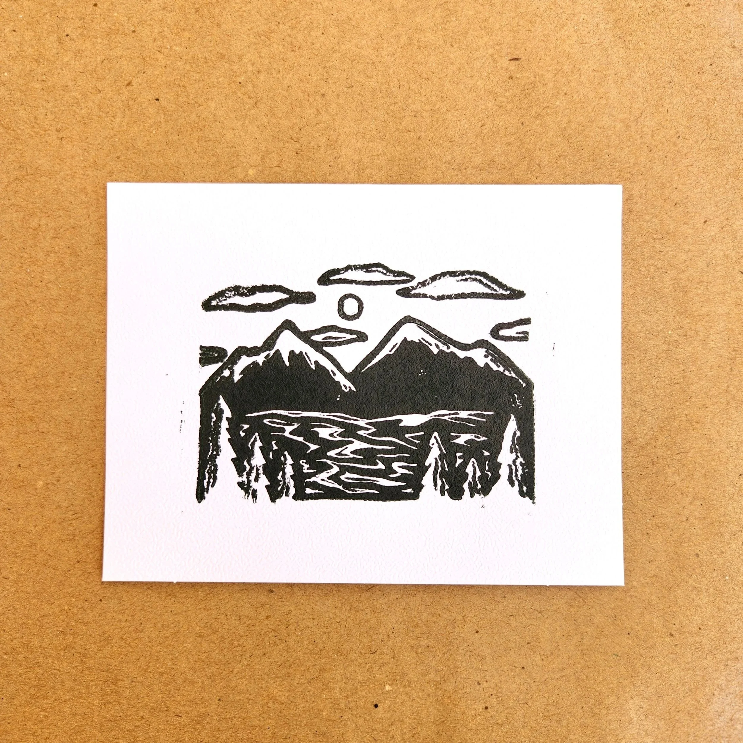 A black and white illustration of mountains, trees, a river, clouds, and a sun on a white piece of paper.