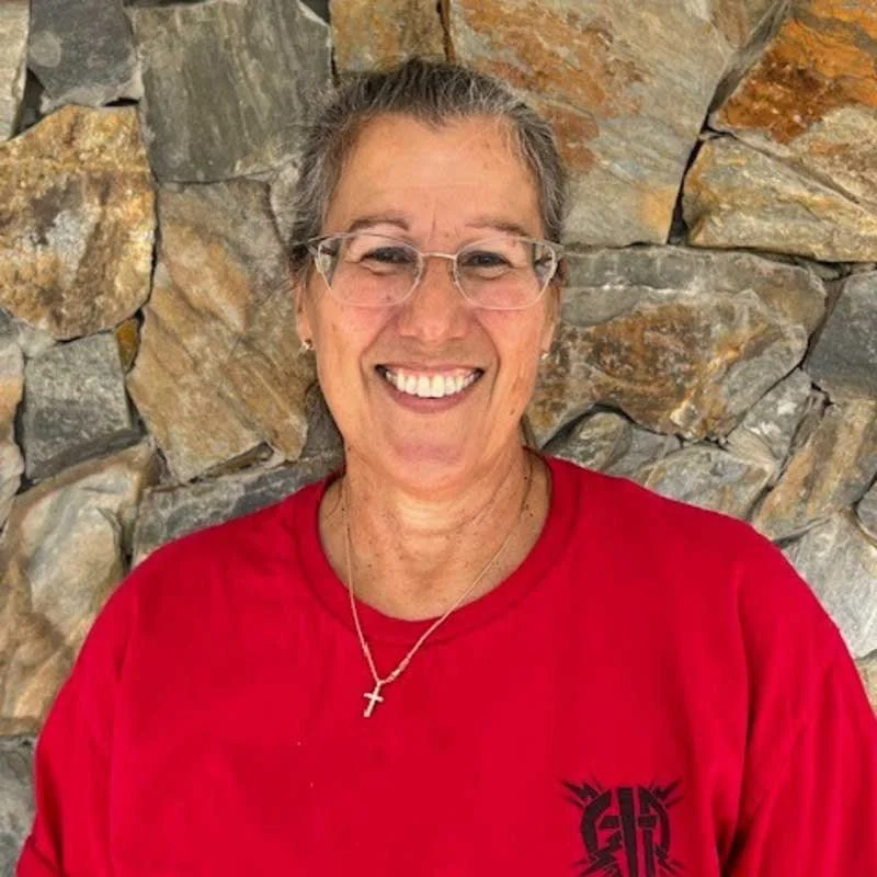 Portrait of Michele Olmstead, Christ In Action headquarters volunteer, smiling in a red shirt against a stone background.