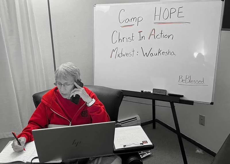 Christ In Action team member in a red sweatshirt taking notes at a desk during a ministry event, with a whiteboard in the background labeled “Camp Hope.”