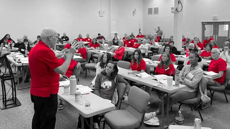 Instructor in a red shirt teaching a Christ In Action training session to a classroom of volunteers seated at tables.