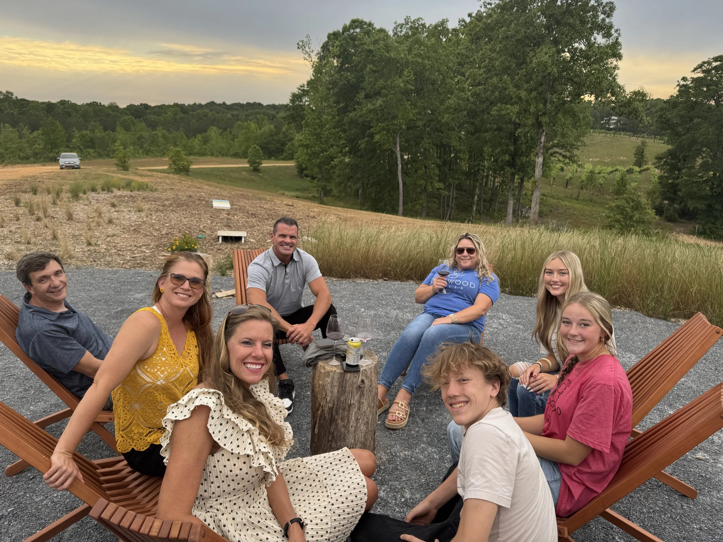 Group of nine people sitting outdoors around a tree, enjoying drinks and smiling, with a rural landscape and cloudy sky in the background.