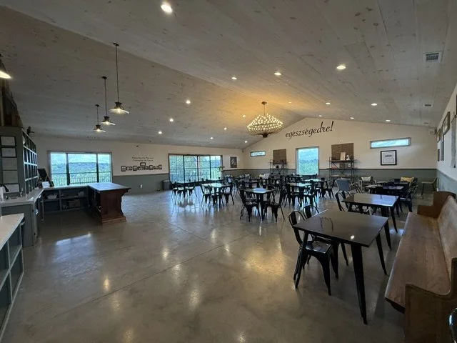 Empty restaurant or cafe with tables and chairs, large windows, pendant lighting, and a chandelier, with a sign on the wall that says 'egészségedre!'