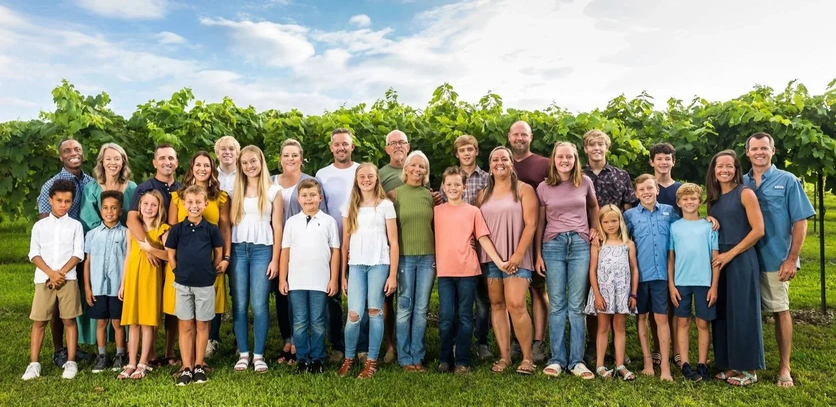 A large, diverse family group standing outdoors in a vineyard with green vines and a partly cloudy sky in the background.