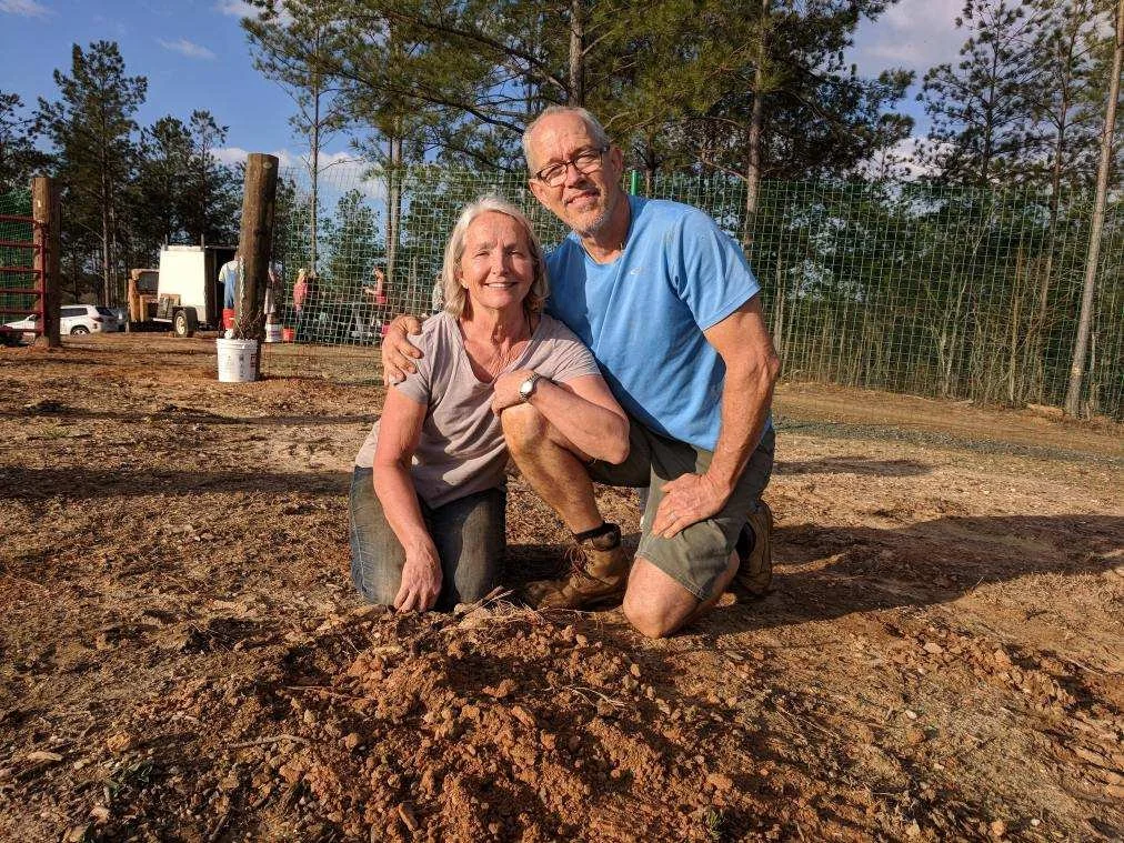 A smiling elderly woman and a smiling elderly man kneeling on the ground outdoors, holding a young tree sapling planted in freshly dug soil, with a background of trees, a fence, and a few people and vehicles in the distance.