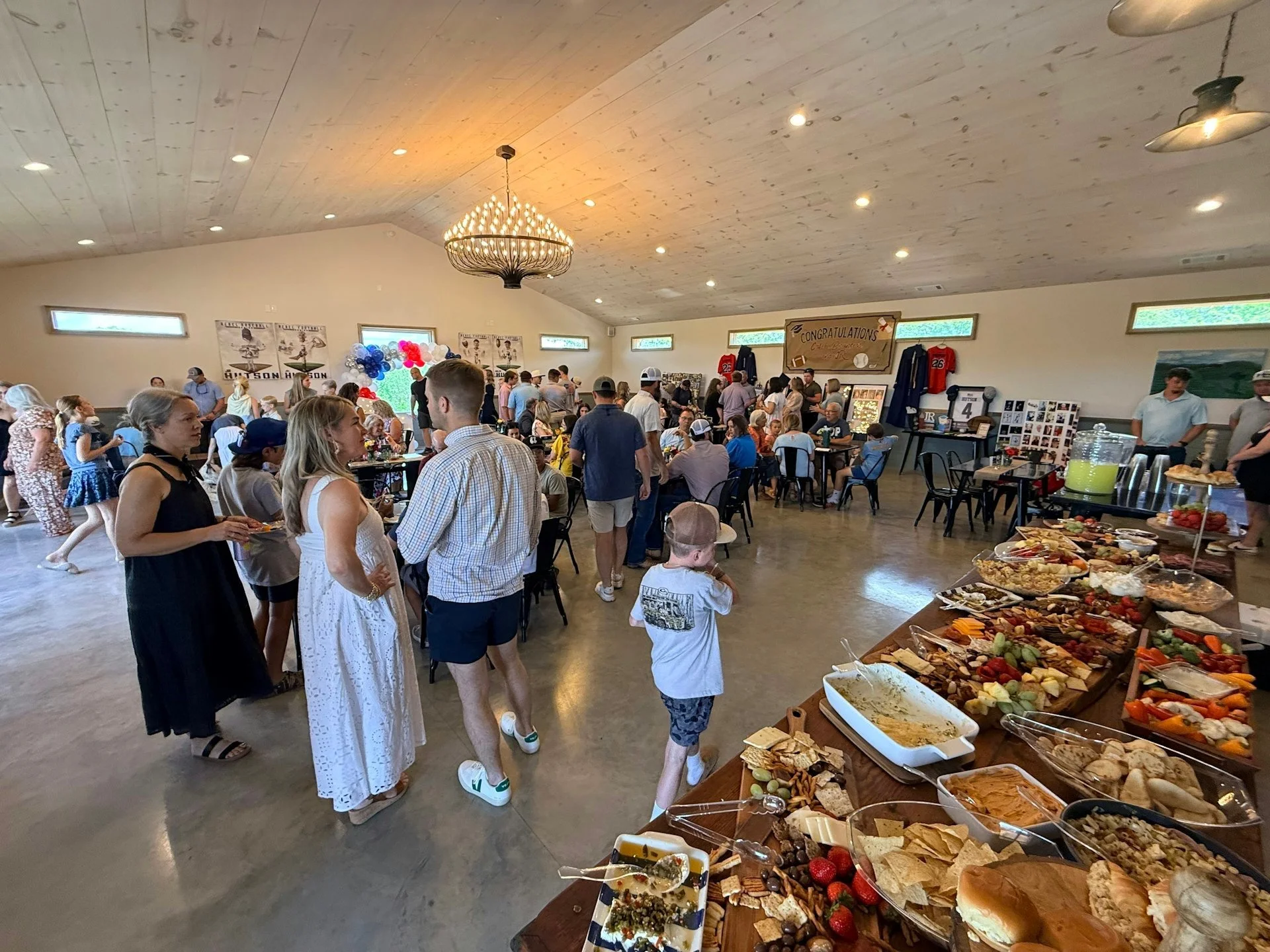 People gathered in a spacious room celebrating an event, with a buffet table of snacks and desserts on the right, and a banner that says 'Congratulations' in the background.