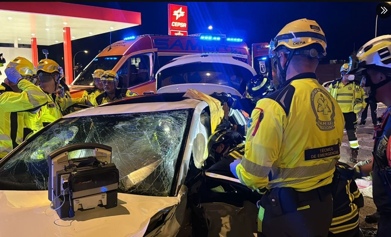 Personal de emergencia atendiendo a víctimas en un accidente de autos en la noche, con un vehículo dañado y una ambulancia en el fondo.