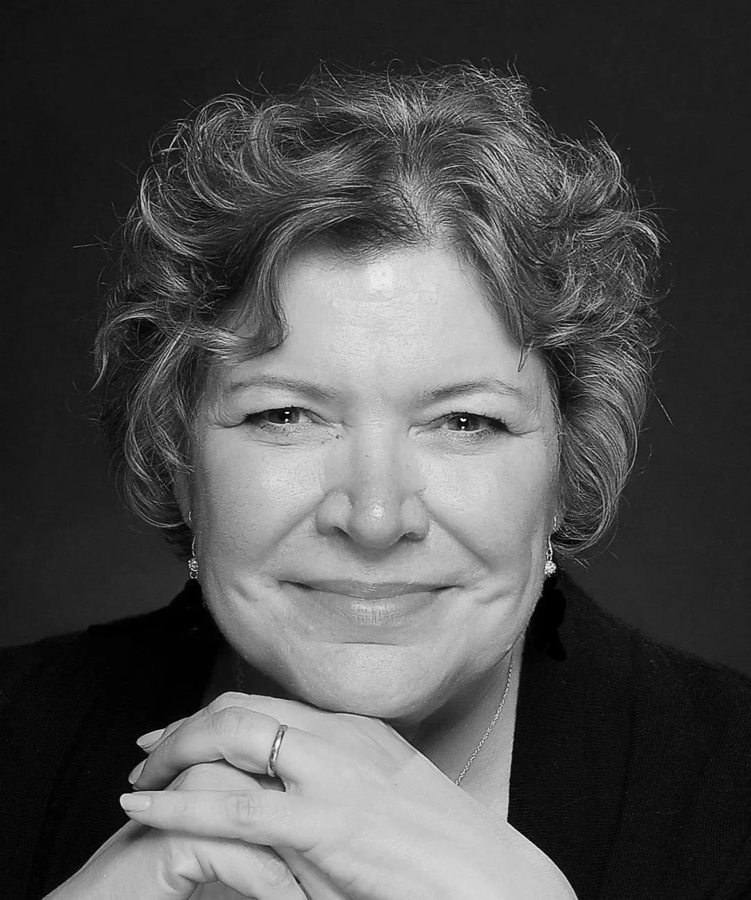 Black and white portrait of a smiling middle-aged woman with short, curly hair, wearing earrings, a ring, and a necklace, against a dark background.