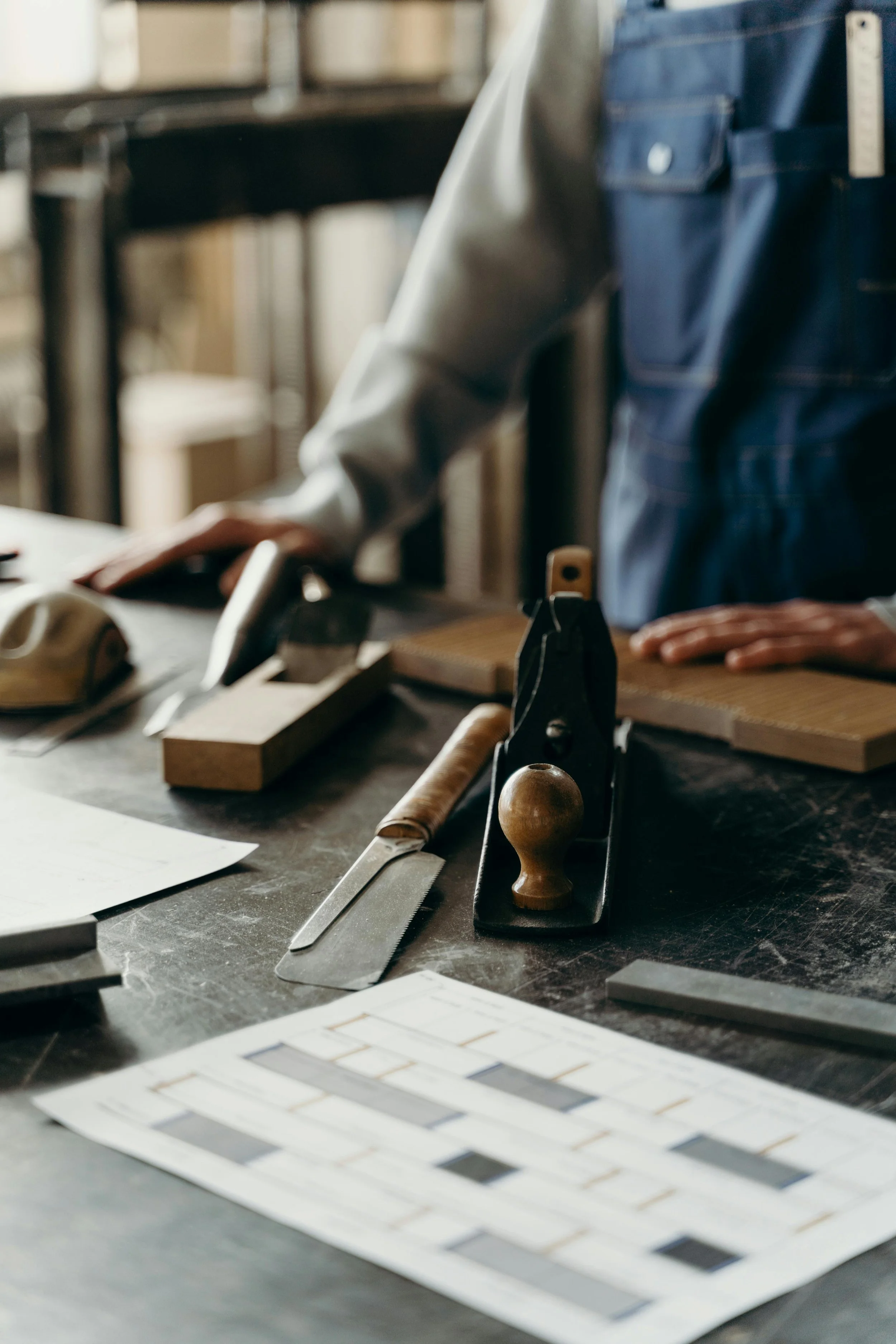 A person working on a woodworking project at a workbench, surrounded by tools such as a hand plane, ruler, and sandpaper, with digital tile samples on the table.