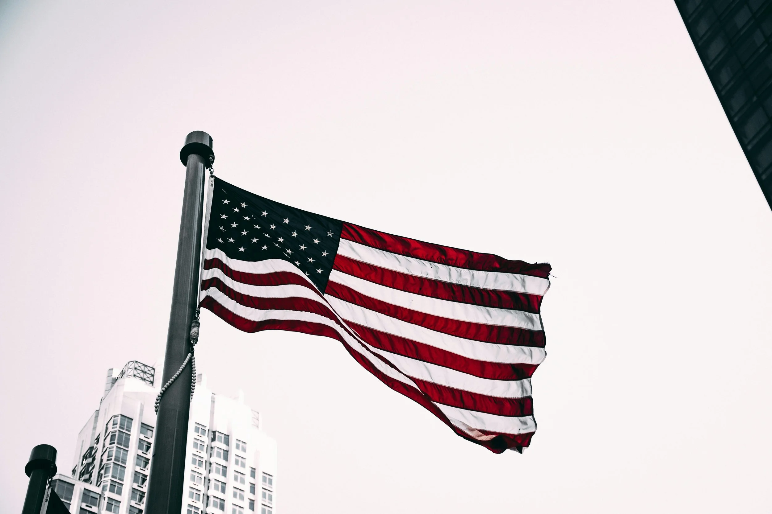 American flag waving in the wind against a gray sky, with tall buildings in the background.