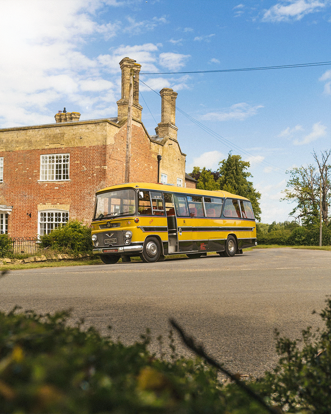 A yellow vintage bus parked on a quiet street in front of a brick building with ivy and greenery, under a partly cloudy sky.