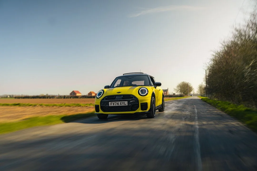 A yellow Mini Cooper car driving on a rural road with green fields and trees on the sides, under a clear sky.