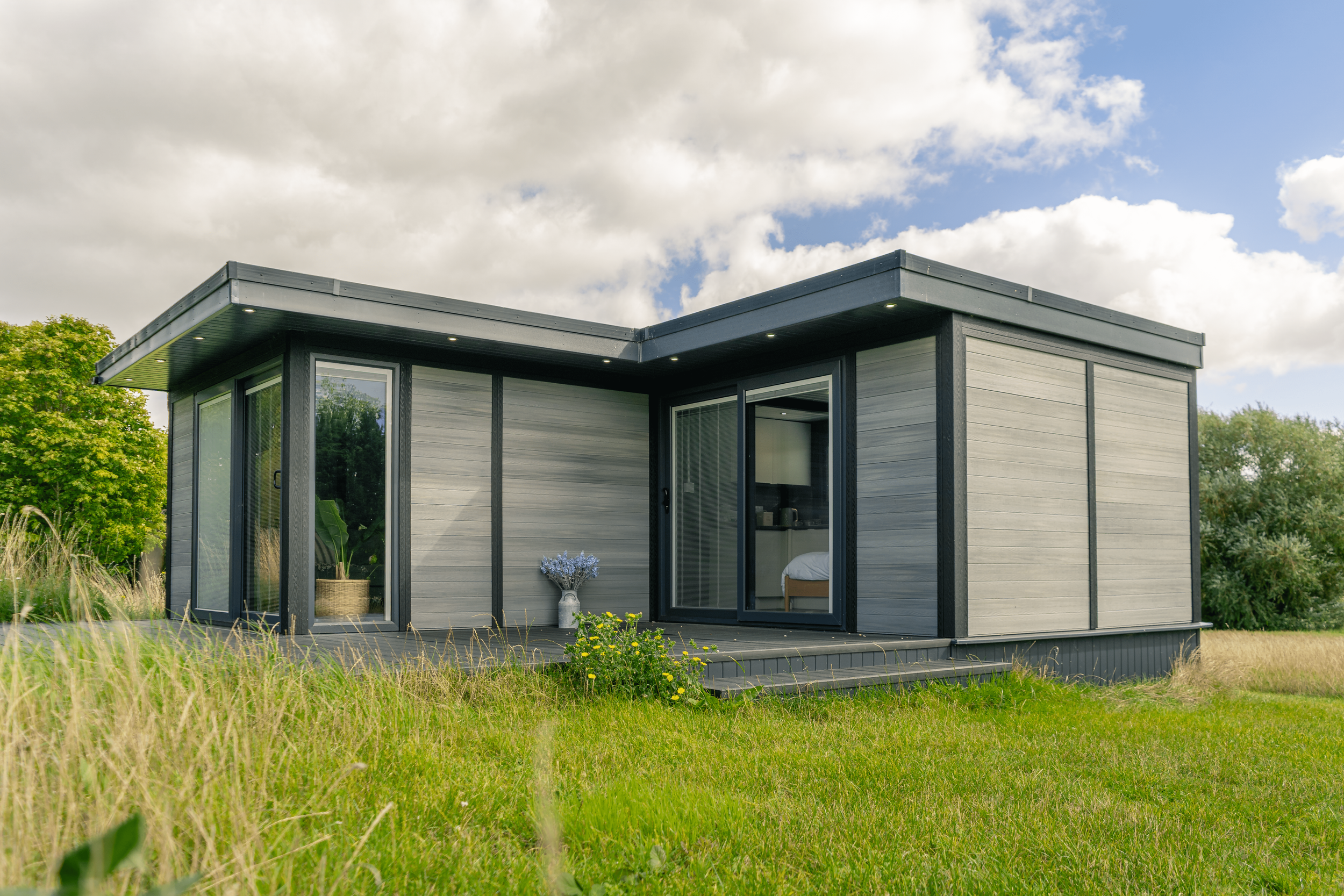 A small modern house with a gray exterior, large sliding glass doors, and a wooden deck, surrounded by green grass and trees under a partly cloudy sky.