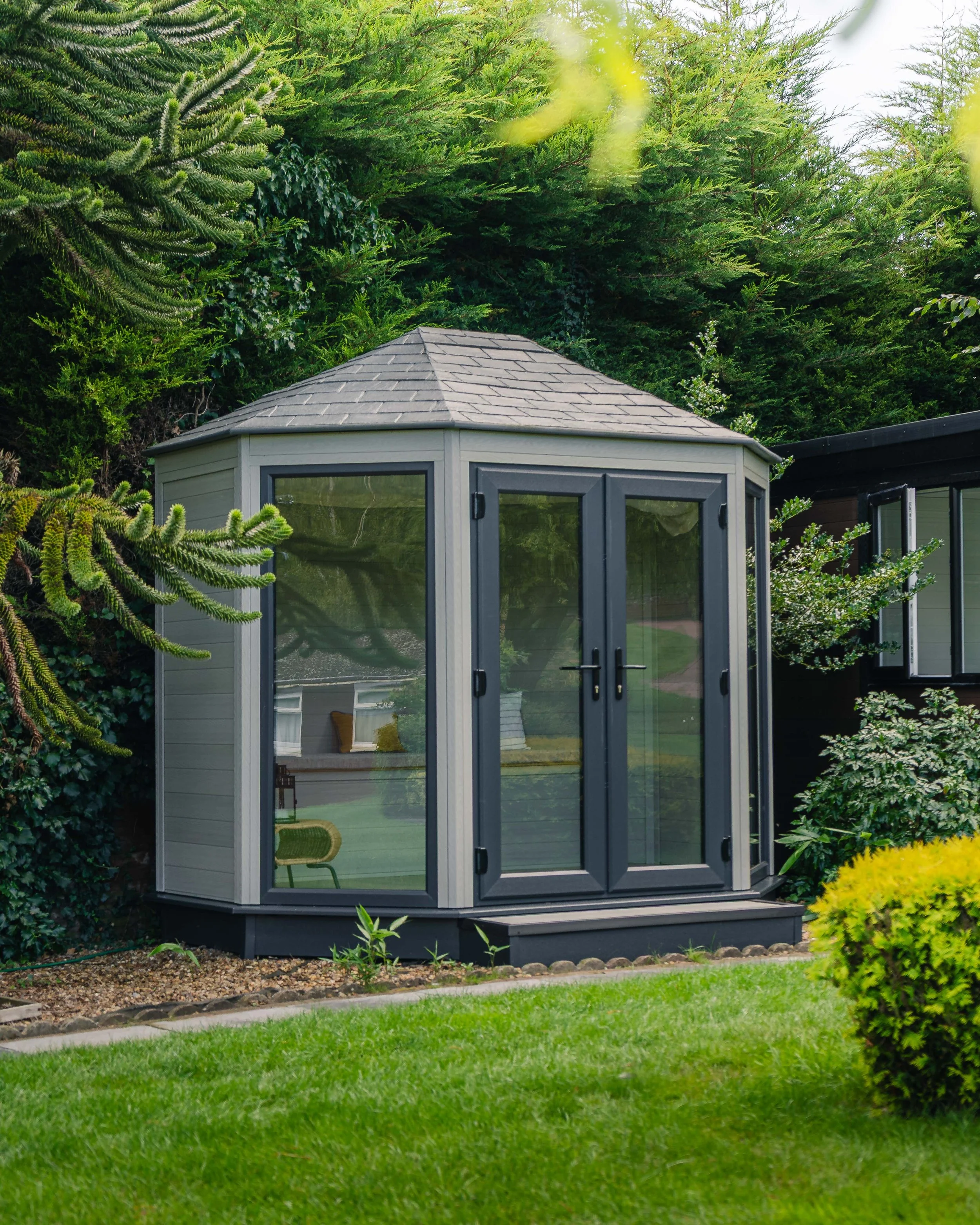A small garden or backyard shed with glass doors, surrounded by lush green plants and trees.