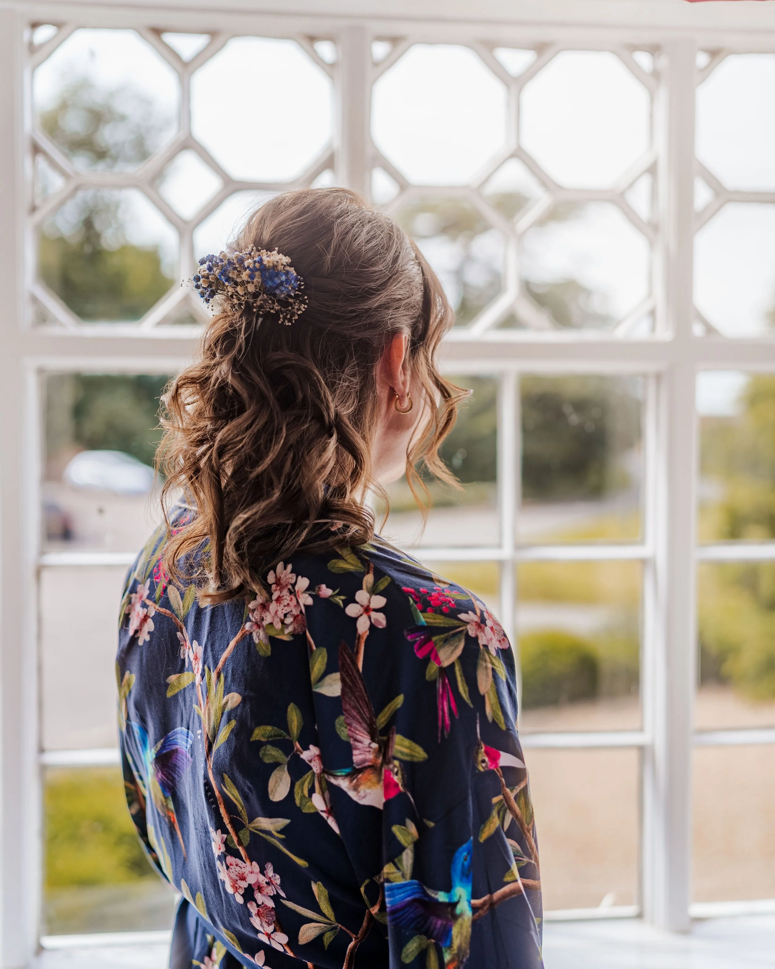 A woman with curly brown hair adorned with a cluster of blue and white flowers, looking out a decorative white window, wearing a dark floral robe with colorful hummingbird prints.