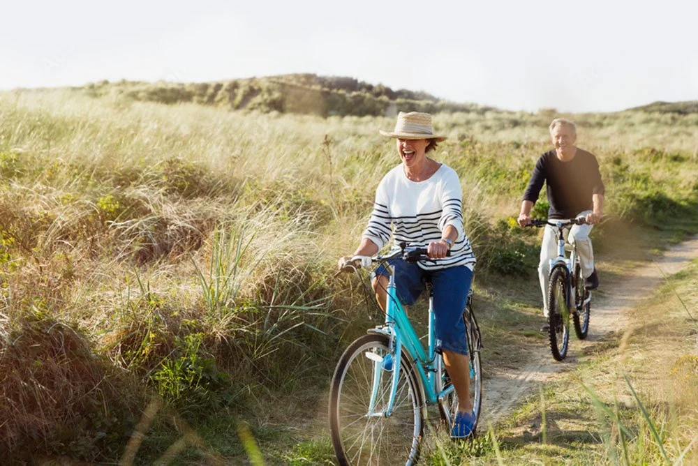 Two retired people cycling on a dirt trail through grassy landscape, one wearing a straw hat and striped shirt, the other in a black shirt, both smiling and enjoying a sunny day.