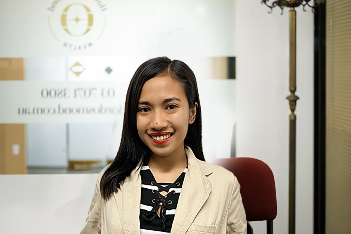 Young woman with long black hair, smiling, sitting in an office environment, wearing a beige blazer over a black and white striped shirt.
