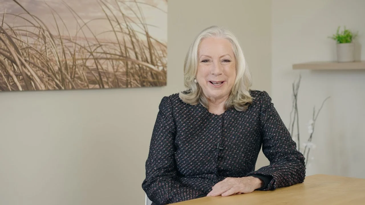 Senior financial planner and principal, Patricia Howard in a blue blazer, with shoulder-length blonde hair smiling, sitting at a wooden table in a room with beige walls, a large piece of abstract art, and a shelf with a potted plant.