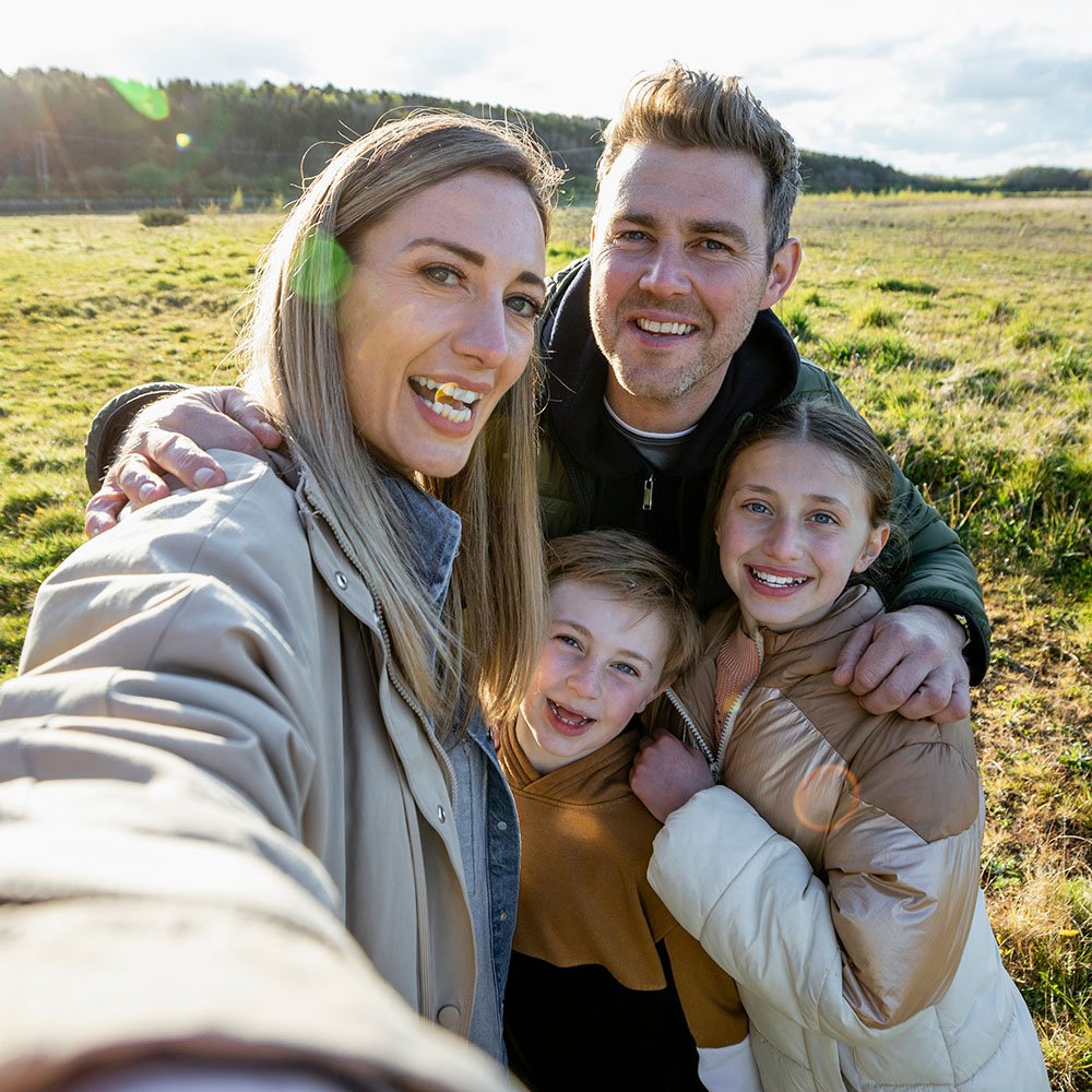 A family of four taking a selfie outdoors in a grassy Melbourne field on a sunny day, smiling and enjoying their time together.