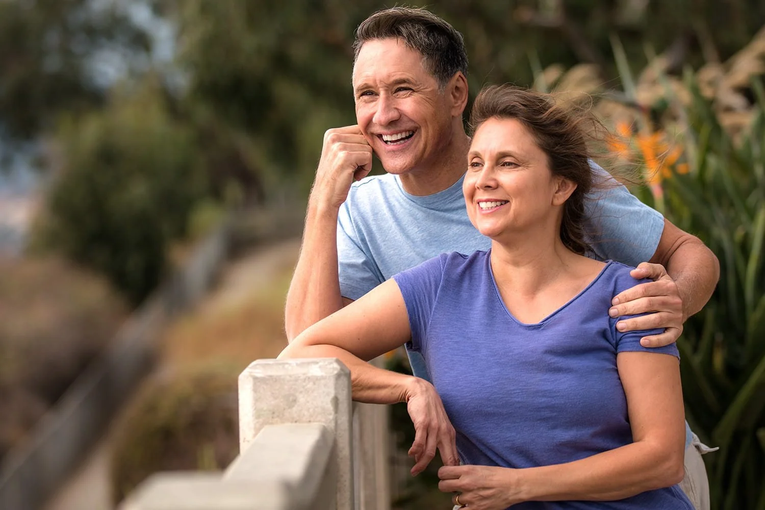 A smiling middle-aged man and woman outdoors, with the man resting his hand on the woman's shoulder, looking into the distance.
