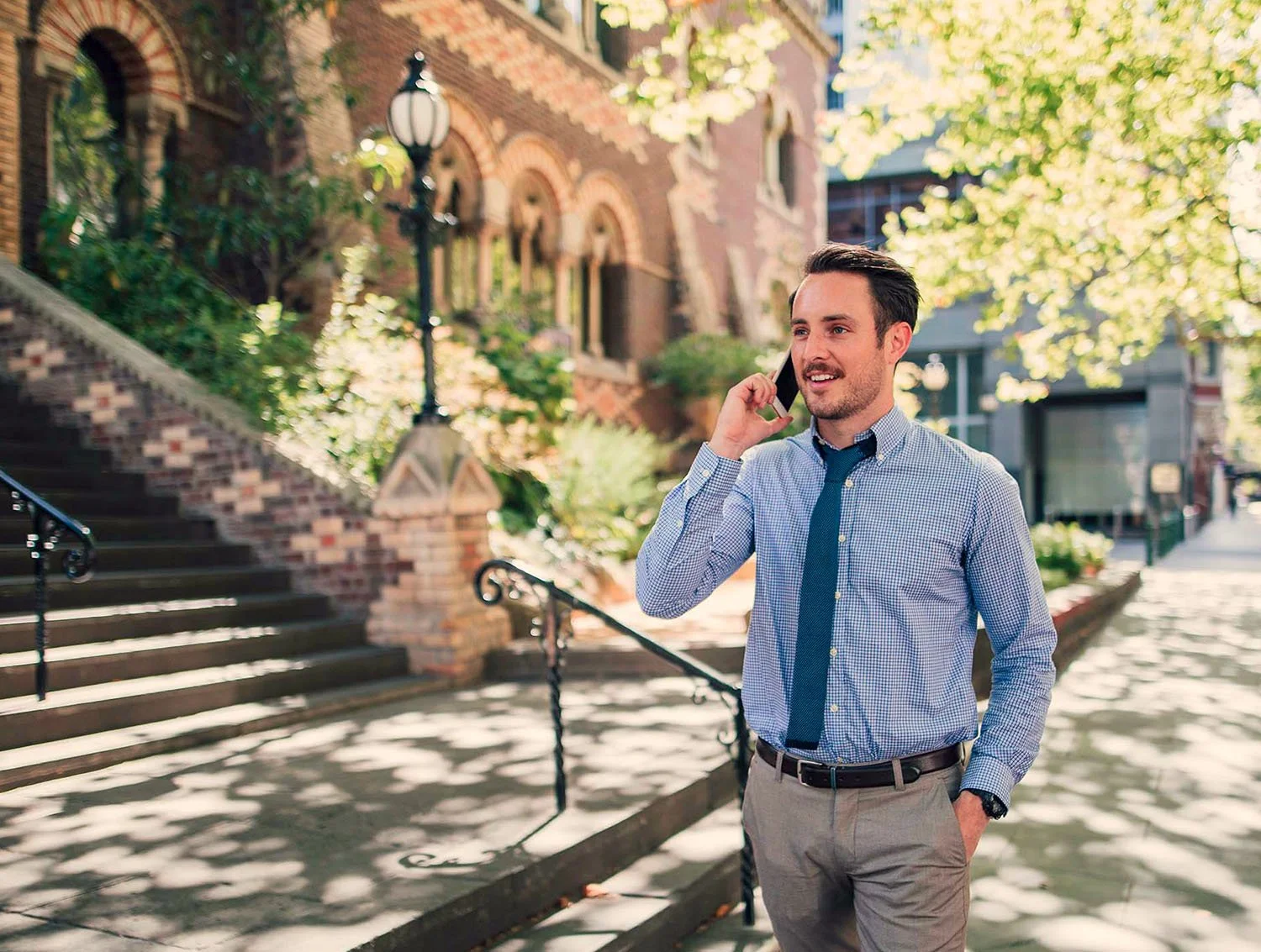 A young man in a blue checkered shirt, tie, and khaki pants talking on his cellphone outdoors on a sunny day, with trees, a brick building, and Melbourne stairs in the background.